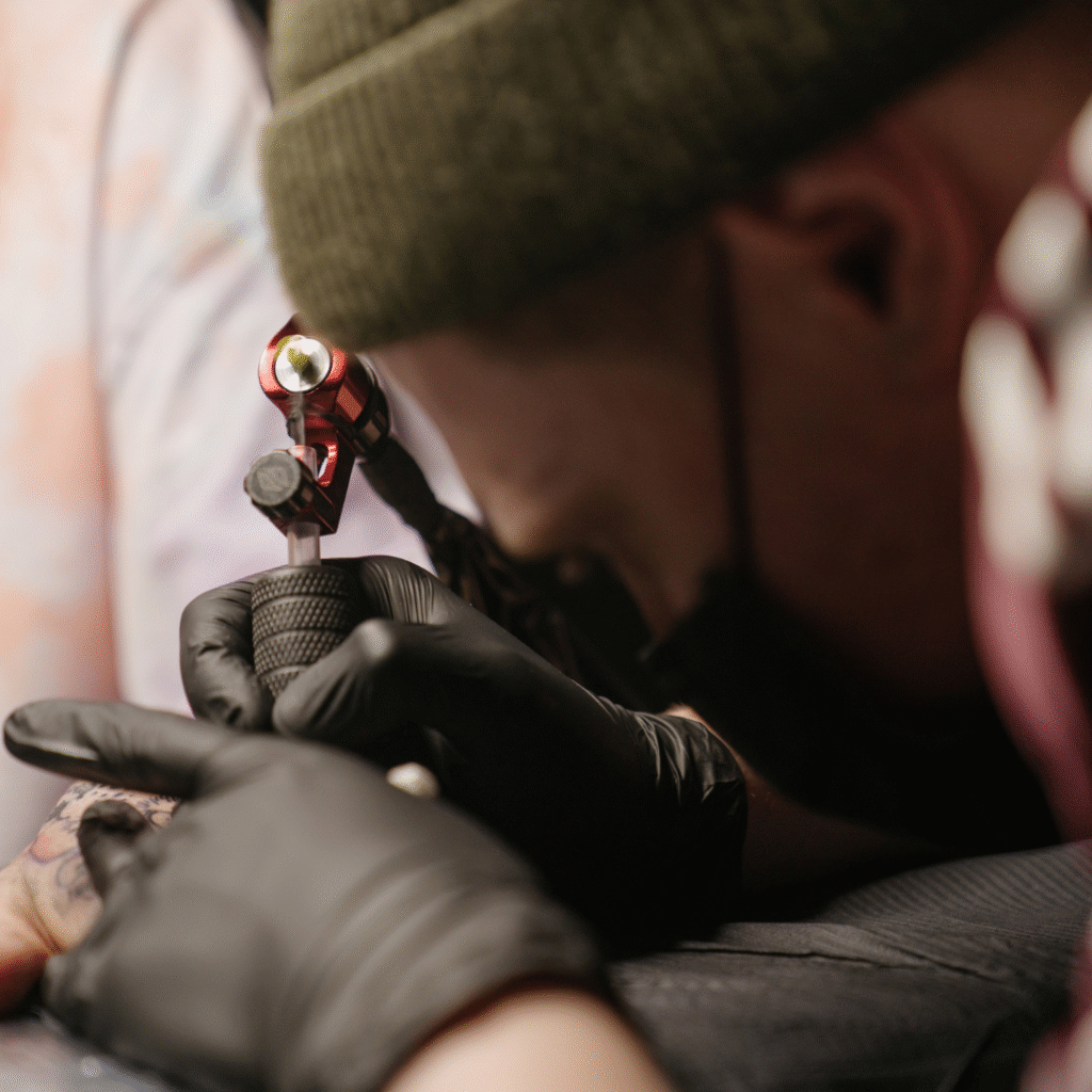A tattoo artist wearing a green beanie and black gloves concentrates while using a tattoo machine on another person's skin. The scene is a close-up, emphasizing the precise technique and intricate details of the tattooing process.