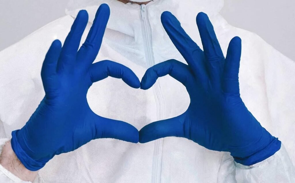 A person dressed in a white protective suit and blue gloves forms a heart shape with their hands, symbolizing compassion and care. The attire and gesture suggest a medical, caregiving, or healthcare setting, conveying warmth and professionalism.
